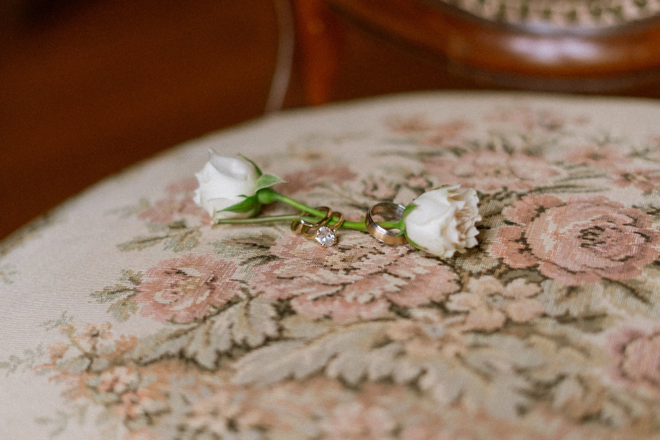 A pair of rings, one with a large clear gemstone, rest on a white flower with green stem and leaves, placed on a decorative fabric surface.