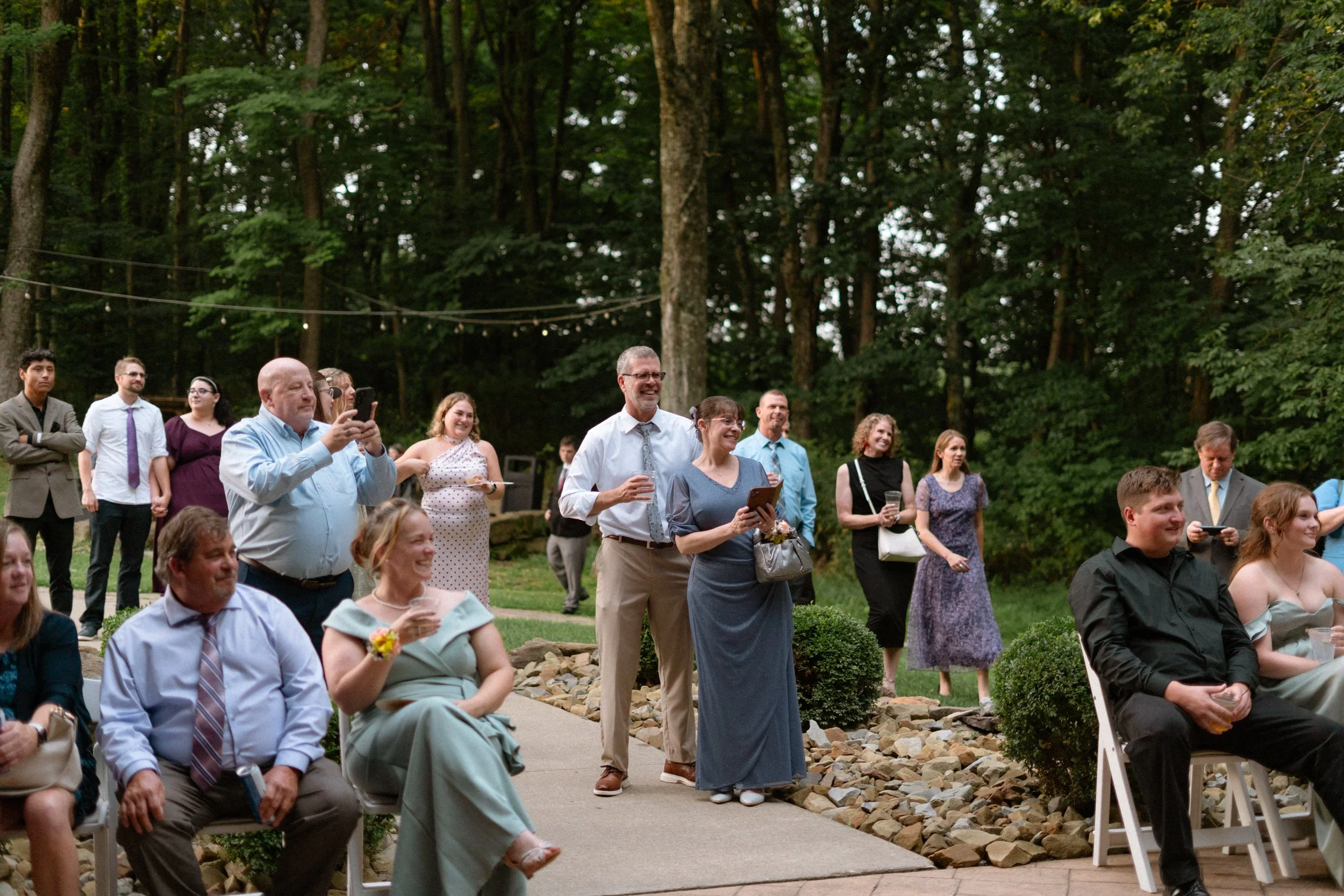 Group of people attending an outdoor event in a wooded area, some standing and some sitting, with string lights overhead and trees in the background.