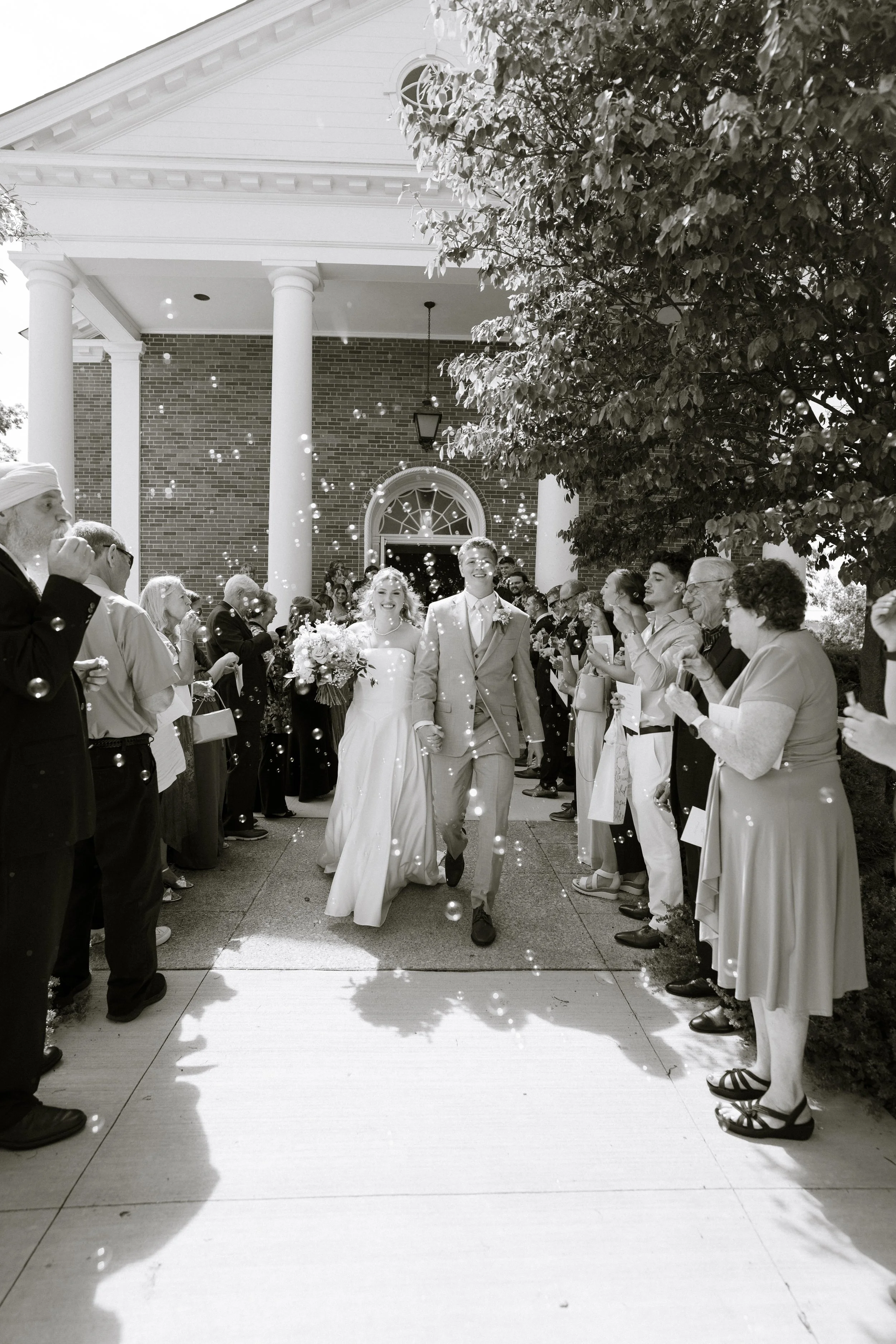 Black and white photo of a bride and groom walking out of a church after a wedding ceremony, surrounded by guests throwing flower petals.