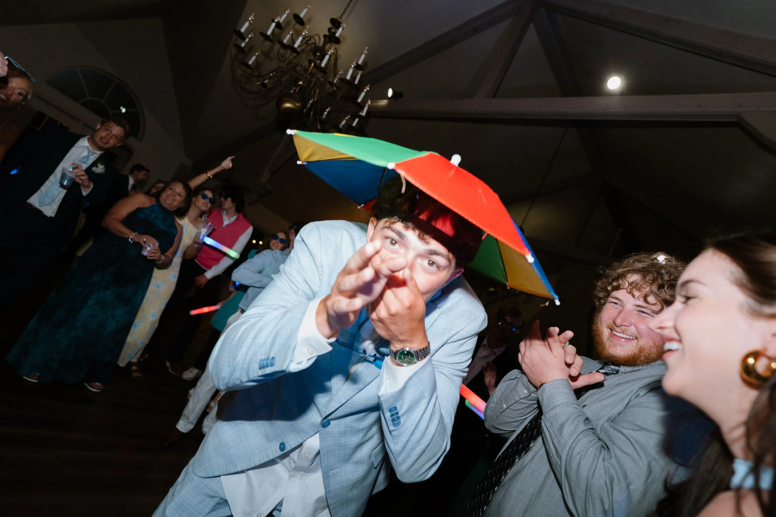 A group of people at a party or celebration dancing and having fun, with many wearing colorful accessories and an umbrella.