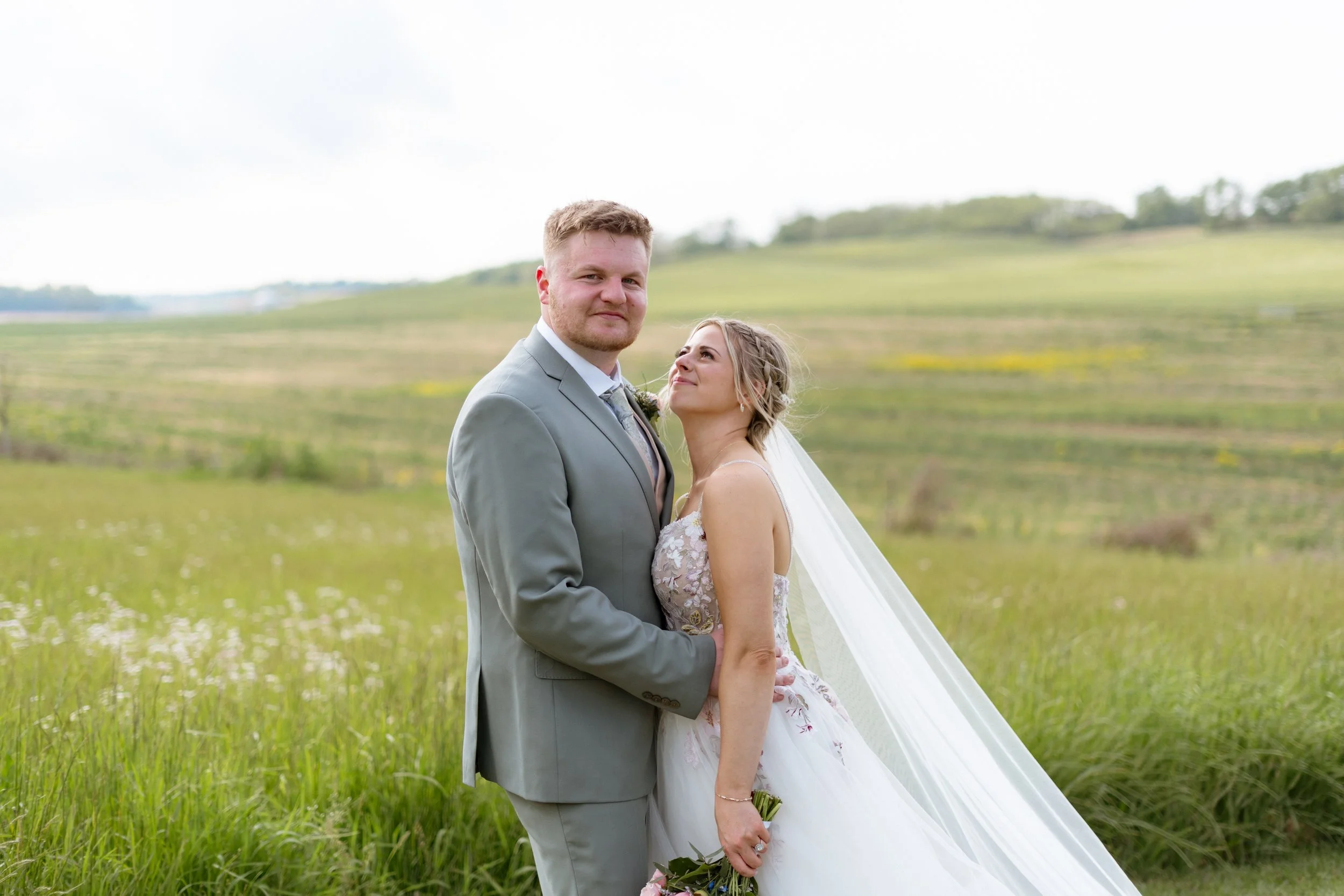 A bride and groom standing together in a grassy field with rolling hills in the background. The groom is wearing a gray suit and the bride is in a white wedding dress with a veil. They are looking at the camera, with the bride smiling and the groom having a slight smile.