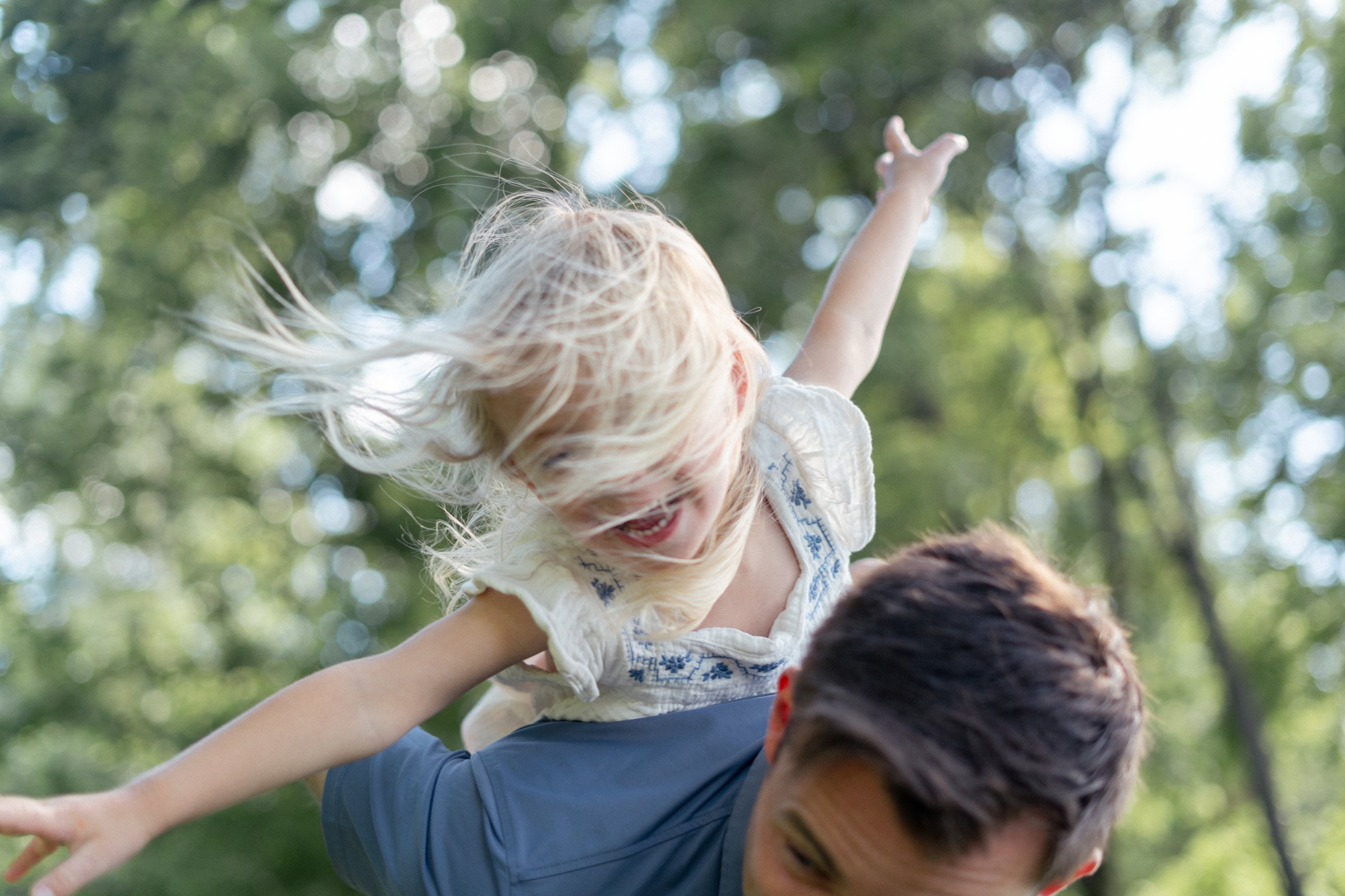A young girl with blonde hair, wearing a white dress with blue embroidery, is sitting on a man's shoulders outdoors in a park with green trees. She is reaching out with her arm, smiling, as they enjoy a sunny day.