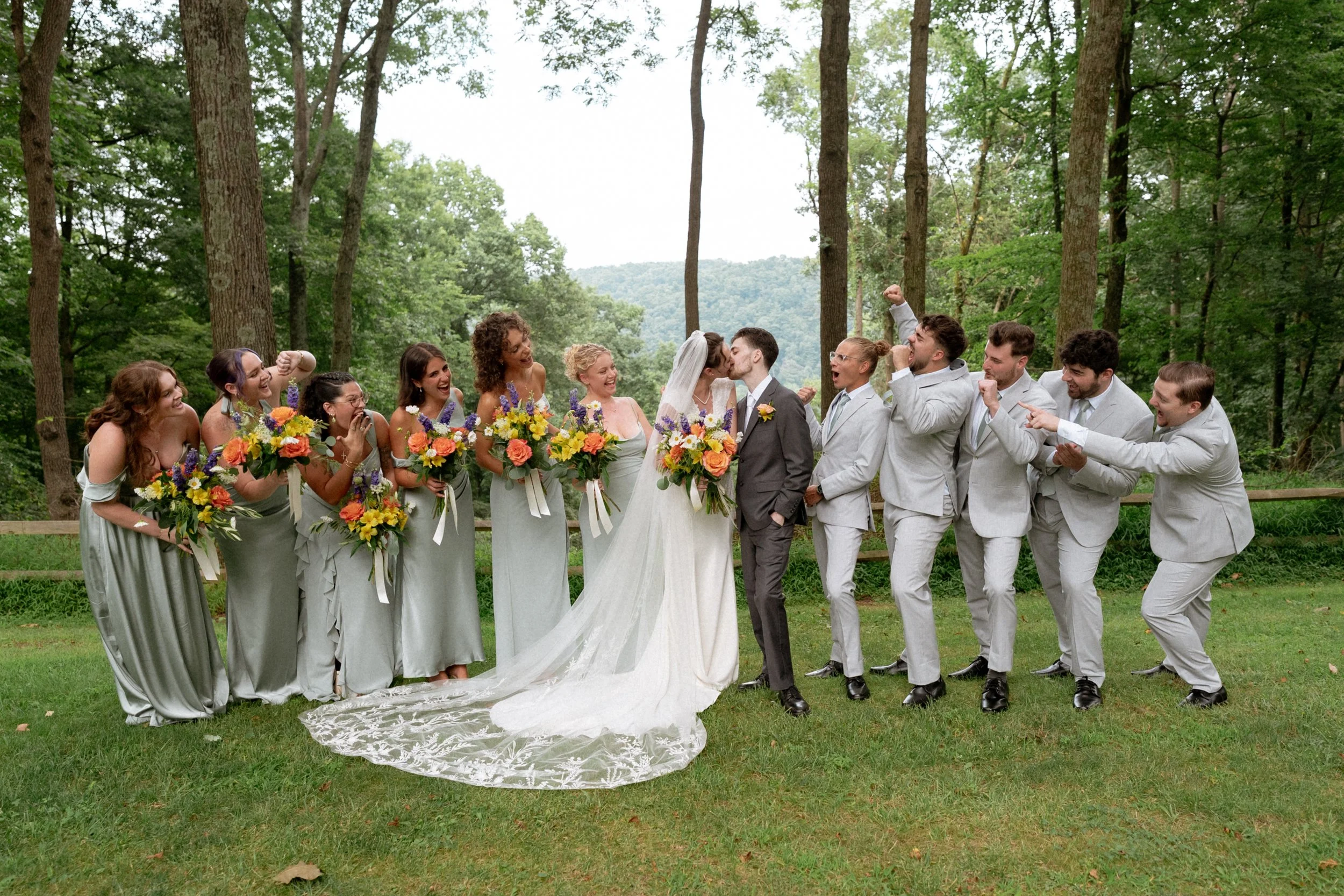 A wedding party standing outdoors in a wooded area, with bridesmaids holding bouquets and the bride and groom kissing in the center.