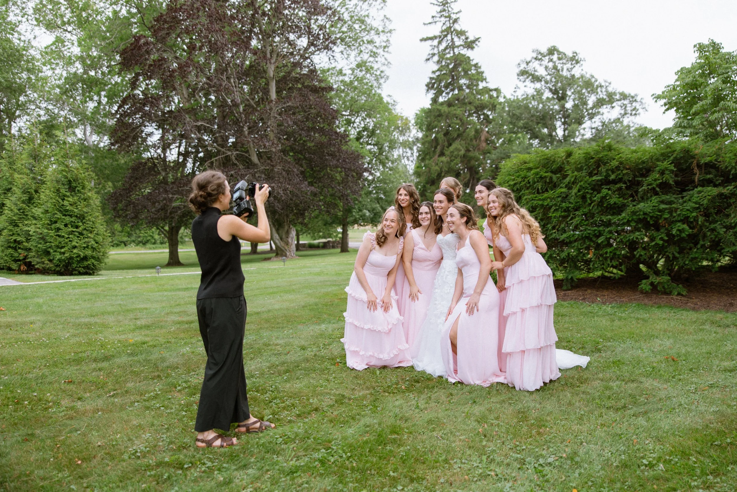 A woman taking a photo of seven women dressed in pink bridesmaid dresses standing outdoors on a lawn with trees in the background.