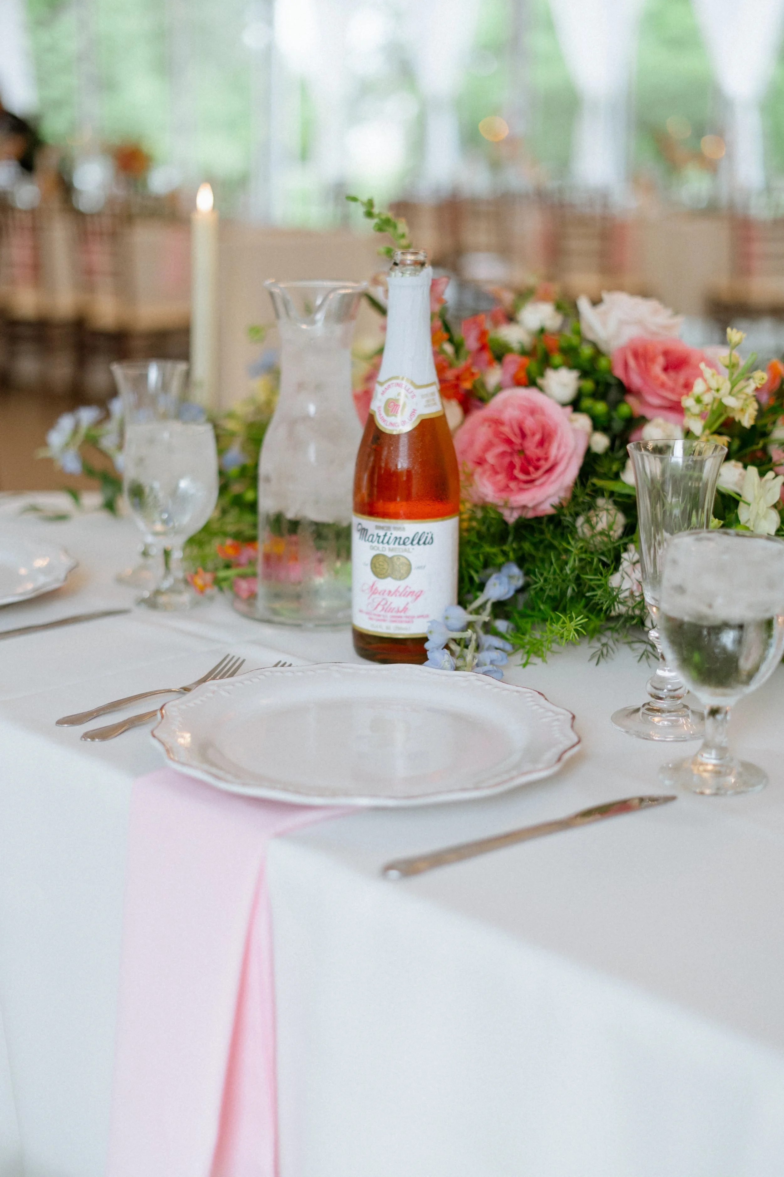 A table set for a celebration with floral centerpieces, a bottle of sparkling blush wine, and elegant glassware, in a room with large windows and natural light.