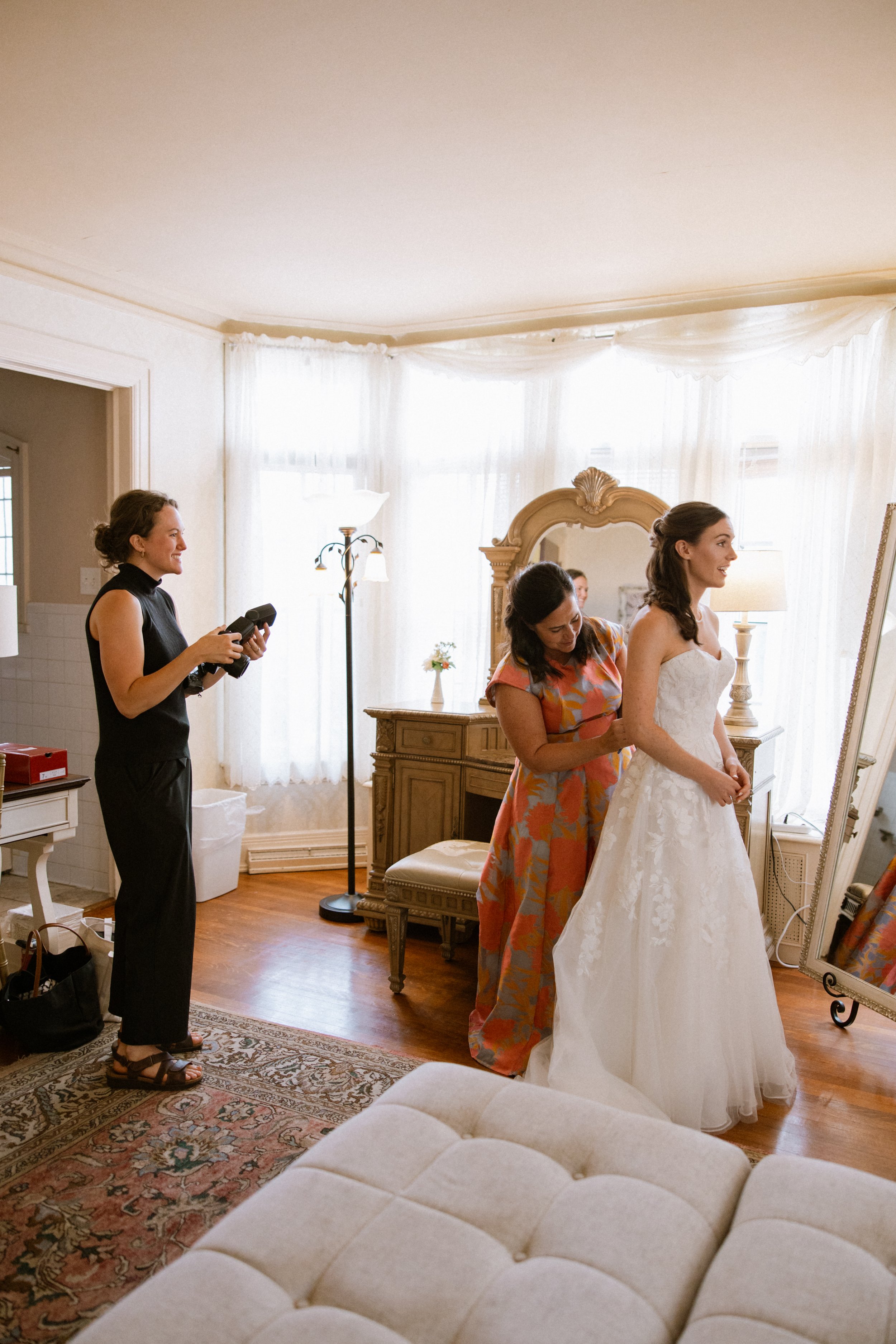 A bride in a white wedding gown is being helped with her dress by a woman in a colorful floral dress. A bridesmaid with dark hair is standing beside her. A photographer in black is capturing the moment. The room has cream-colored walls, white curtains, a wooden dresser, and a full-length mirror. A cushioned ottoman is in the foreground.