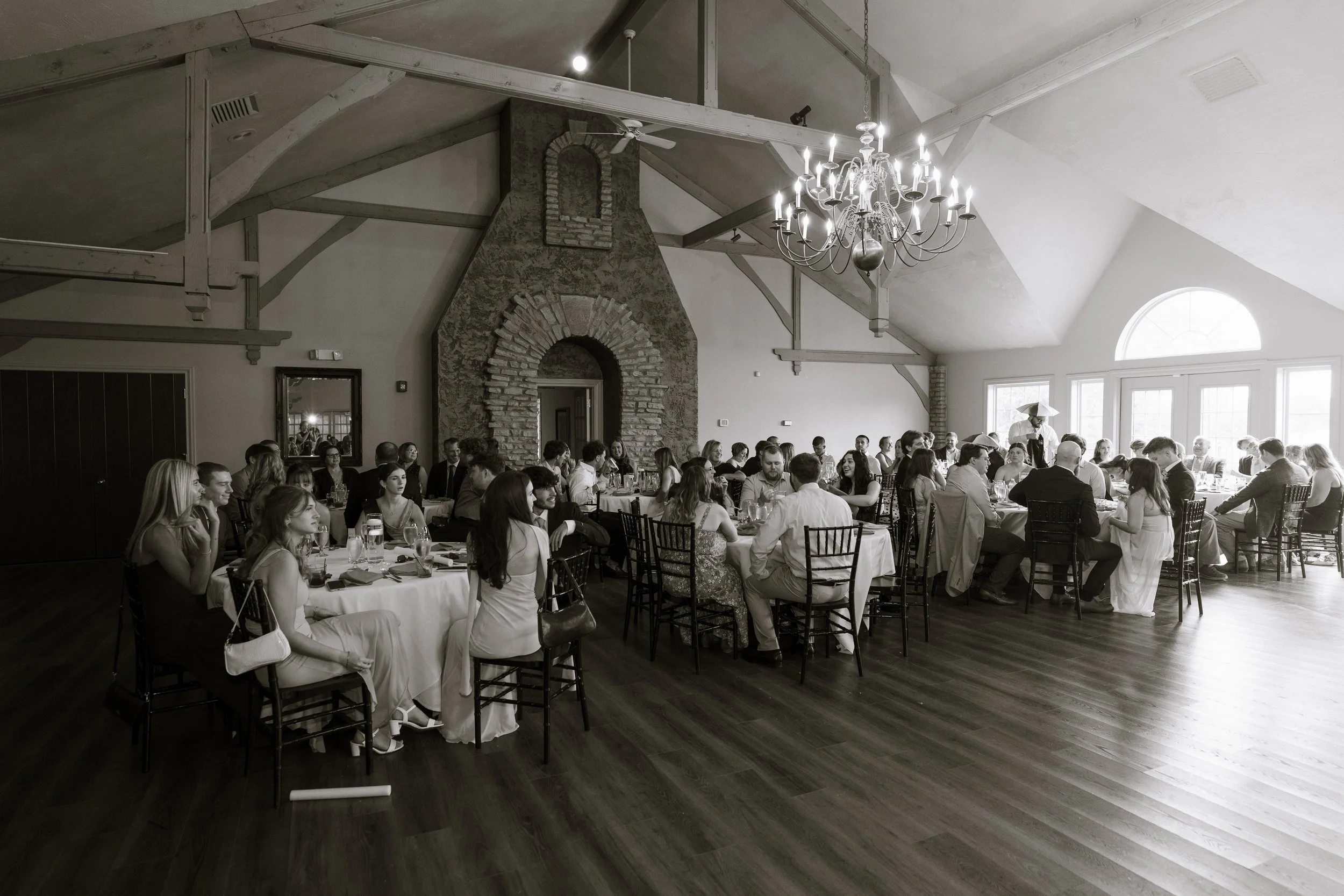 Large indoor gathering of people seated at round tables in a rustic decorated banquet hall, with a large chandelier hanging from the ceiling, white walls, and large windows allowing natural light.