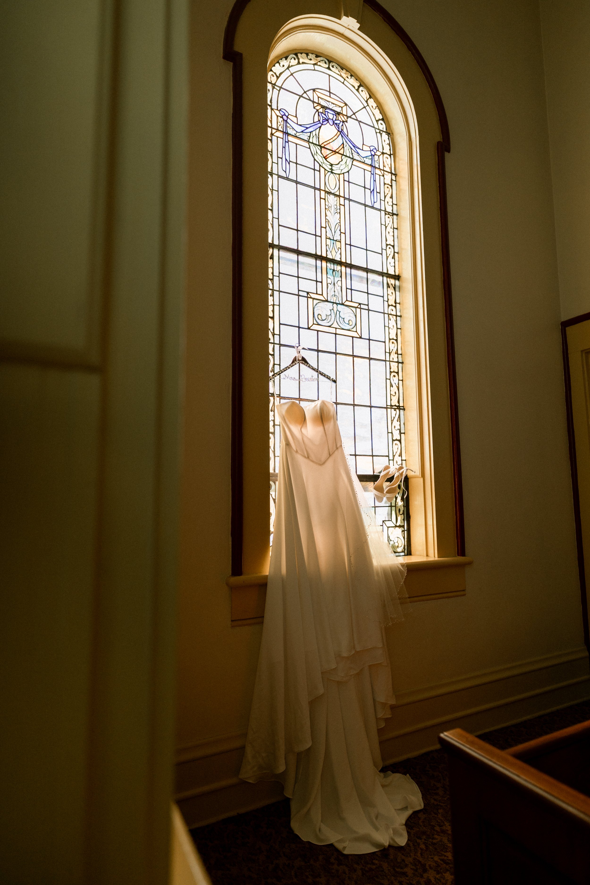 A wedding dress hanging in front of a tall stained glass window in a church or chapel with beige walls and dark trim.