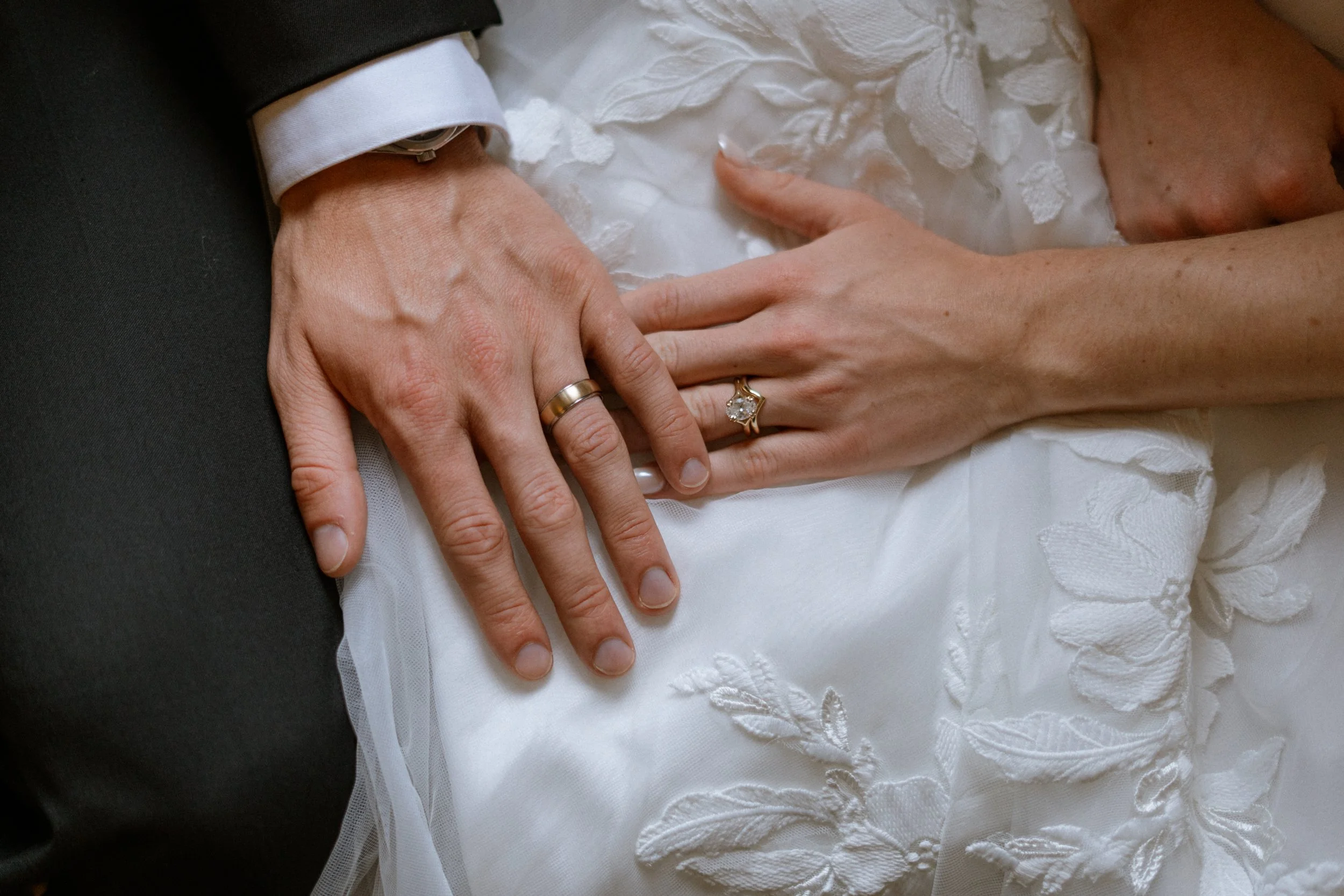 Close-up of a wedding couple's hands, showing wedding rings, with one hand resting on a white textured wedding dress.
