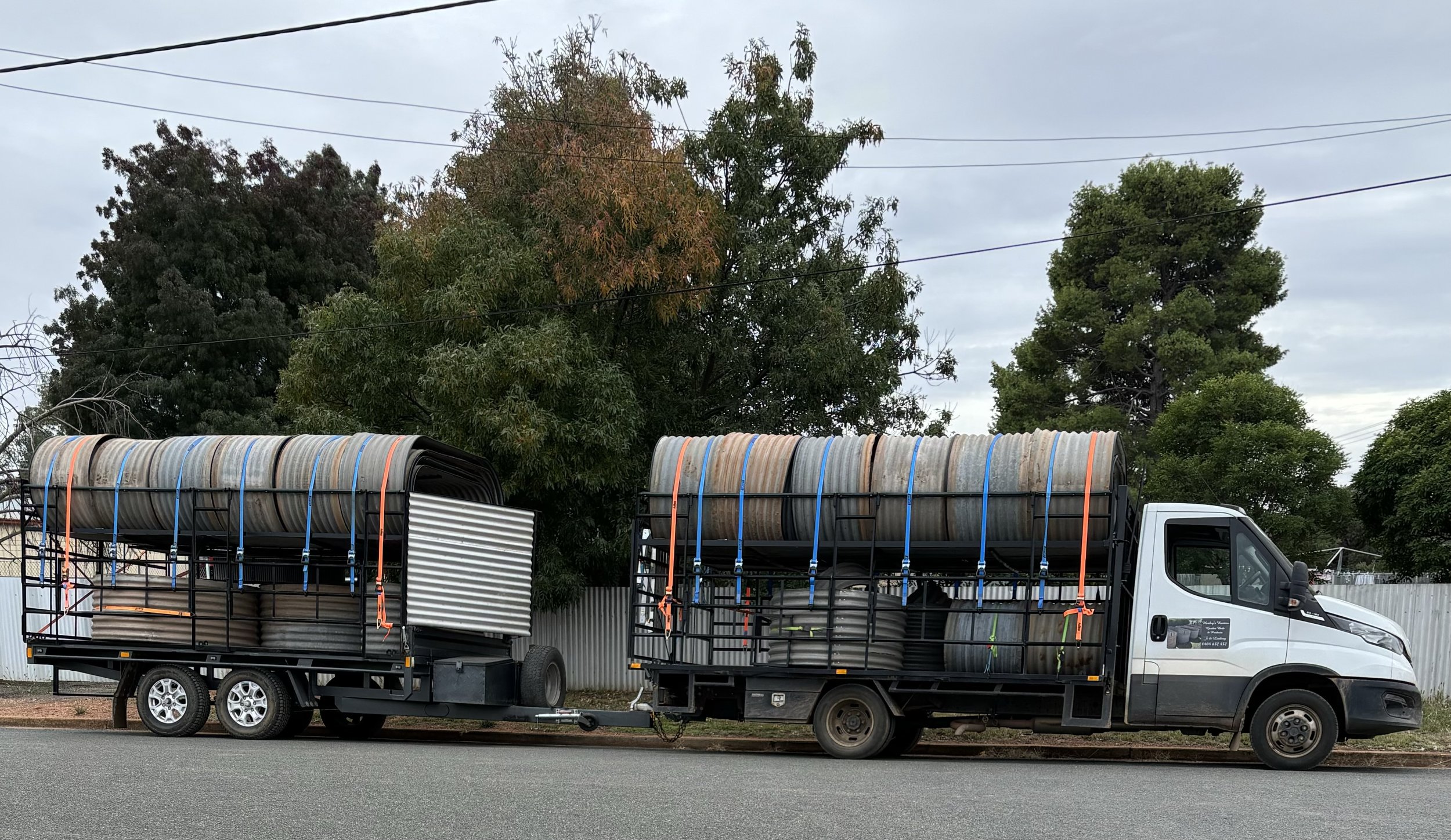 A truck and a trailer parked along the street, both loaded with various pipes and hoses secured with straps, with trees and a cloudy sky in the background.