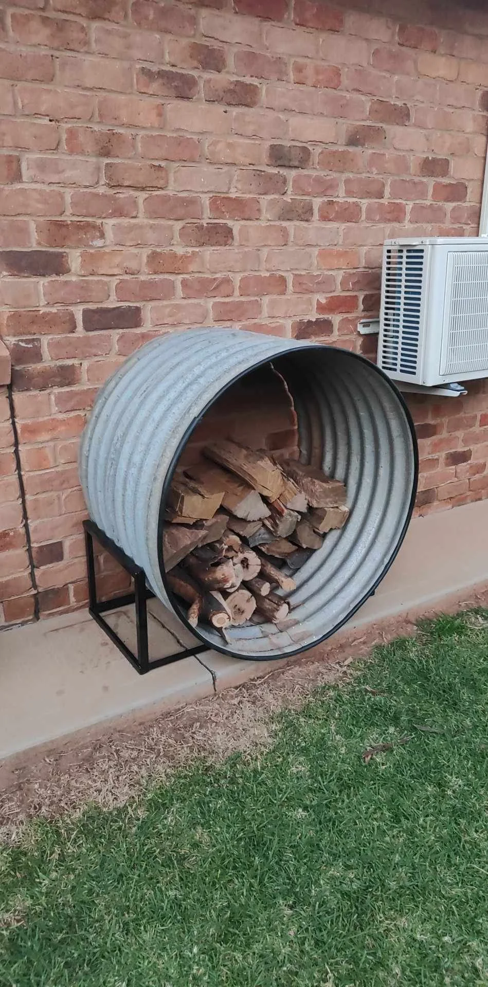 A metal barrel filled with chopped firewood leaning against a brick wall, with an air conditioning unit mounted on the wall nearby.