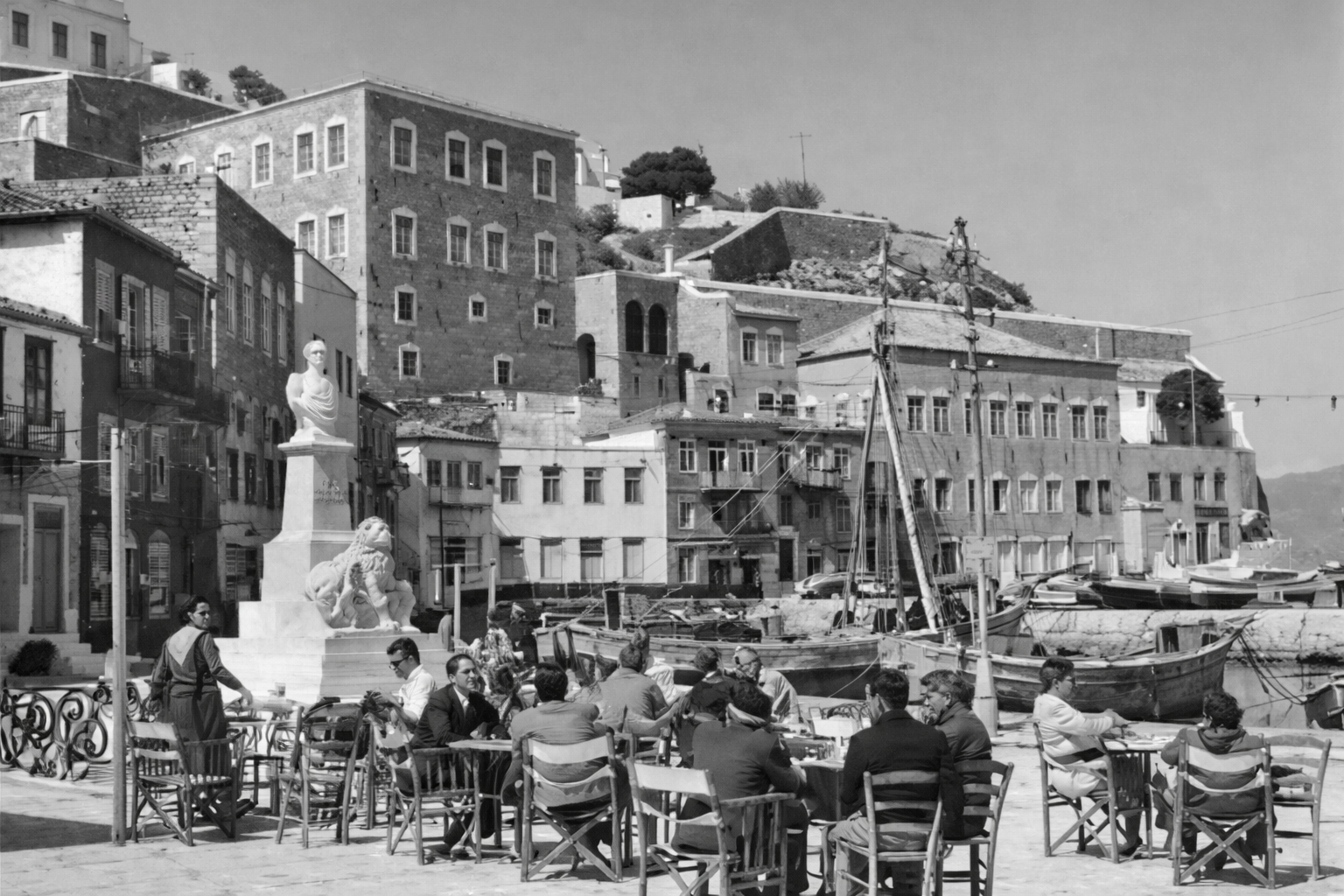 A group of people dining outdoors at a cafe near a marina with boats, in front of a hillside with old buildings and a statue.