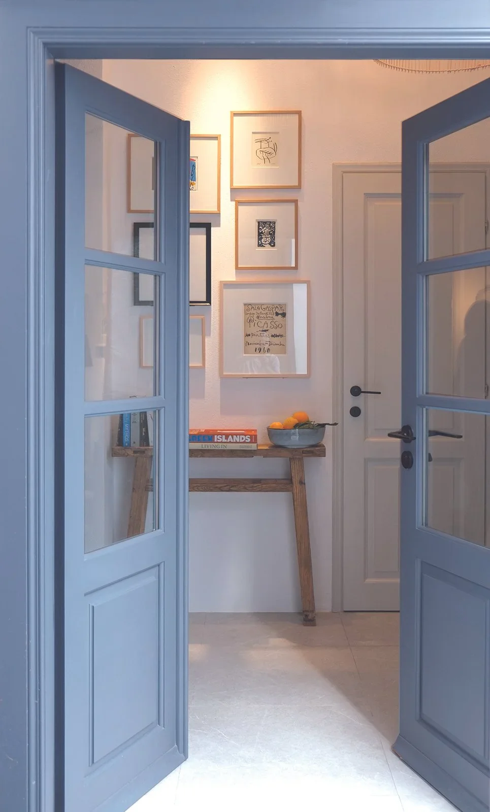 A hallway view through two blue French doors, with a wall decorated with framed artwork, a small wooden console table with books and a bowl of oranges.