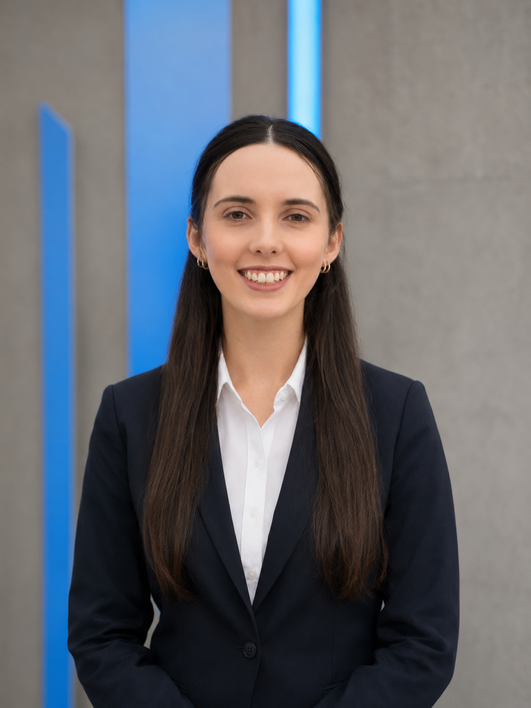 A young woman with long brown hair smiling, dressed in a dark blue jacket, standing in front of a concrete wall with illuminated blue panels.