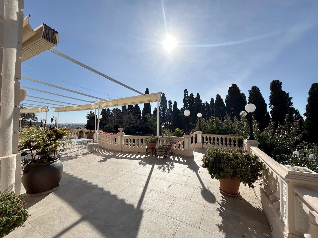 Bright patio with potted plants, a cream-colored balustrade, and a retractable awning under a clear blue sky with the sun shining overhead.