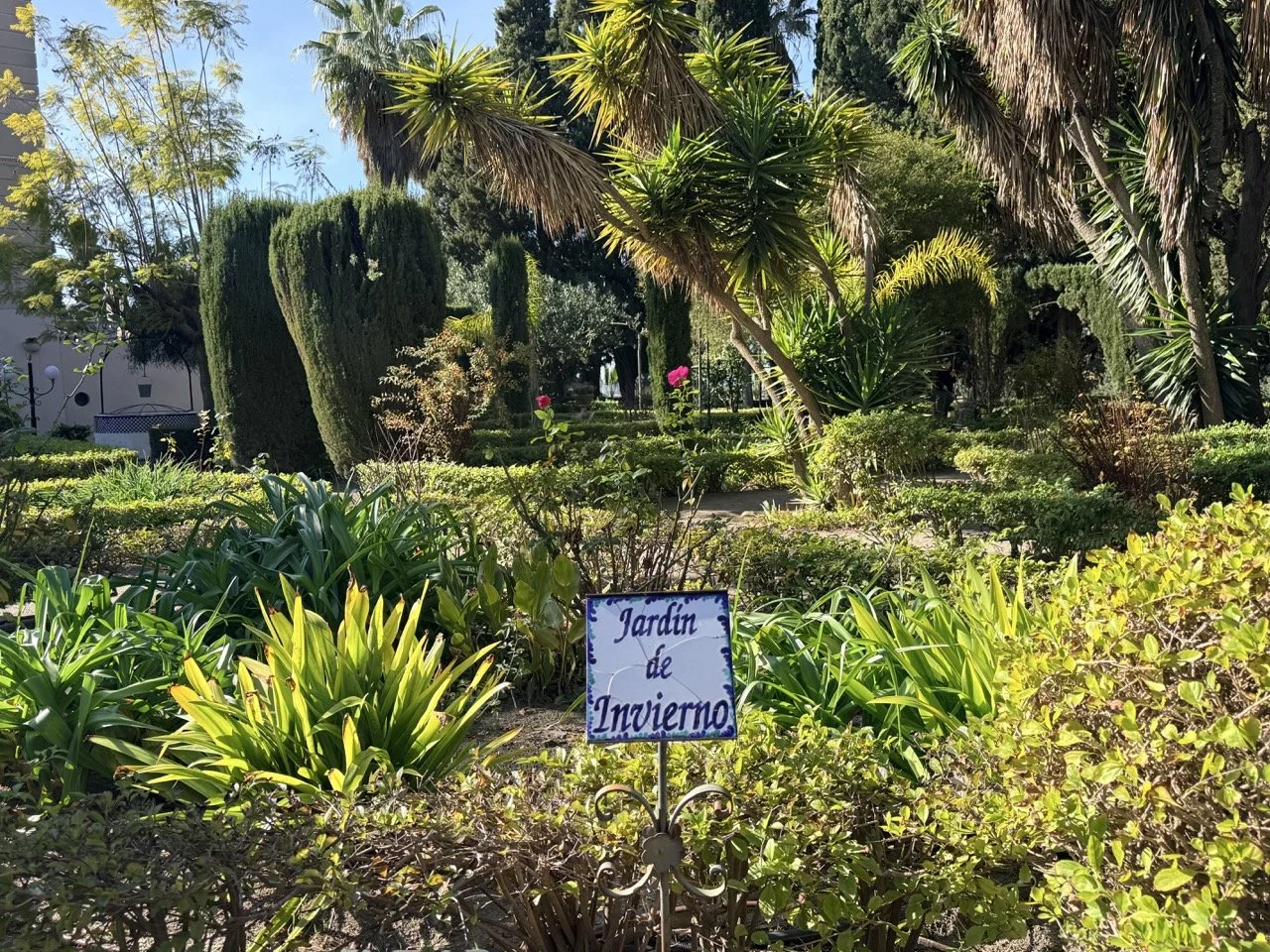 A lush garden with various green plants and trees, including palm trees, with a sign that reads 'Jardin de Invierno' indicating it is a winter garden.