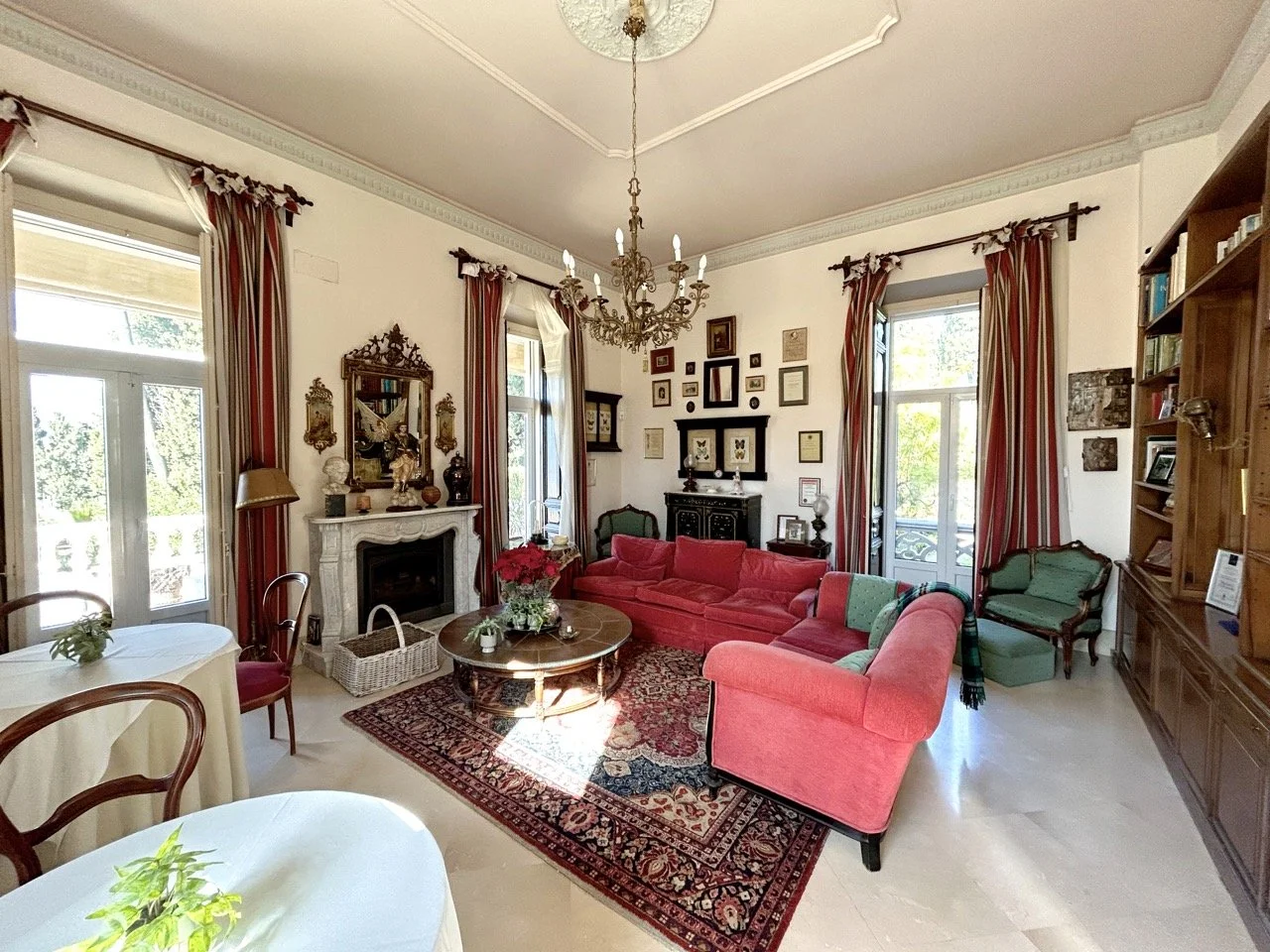 Living room with large windows, red and green upholstered furniture, a fireplace, a chandelier, framed artwork and mirrors on the walls, bookshelf, and a patterned area rug.