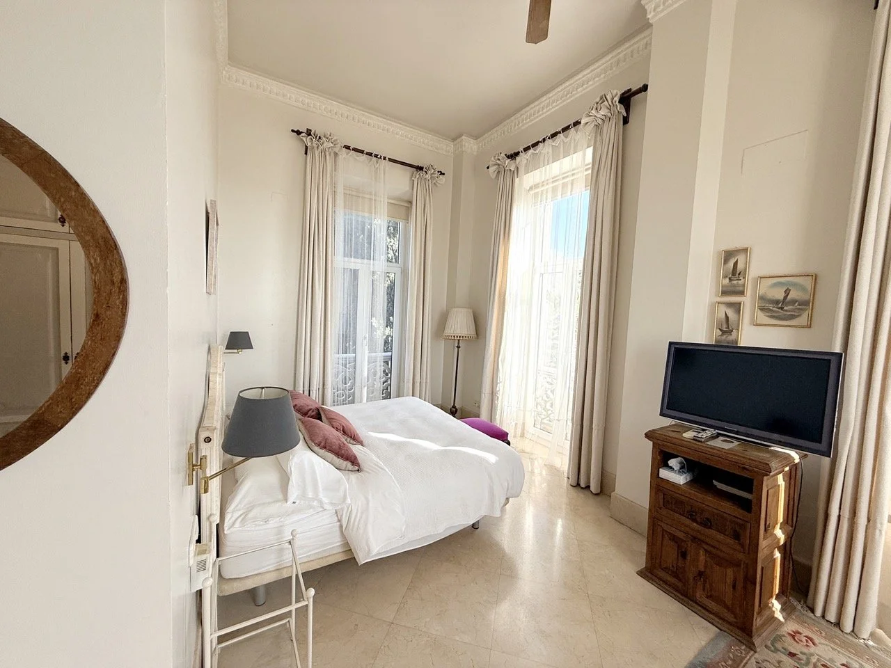 A cozy bedroom with white walls and beige tile flooring, featuring a bed with white bedding and red pillows, a wooden TV stand with a flat-screen TV, and large windows with white curtains allowing natural light.