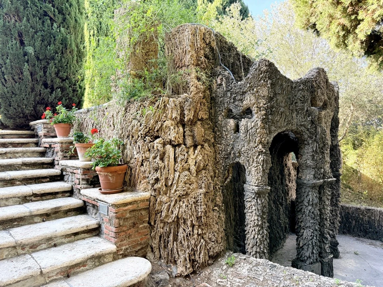 Stone stairs with potted plants and a large, textured tree stump that appears to be a carved animal, possibly a bear, in a garden surrounded by trees and greenery.