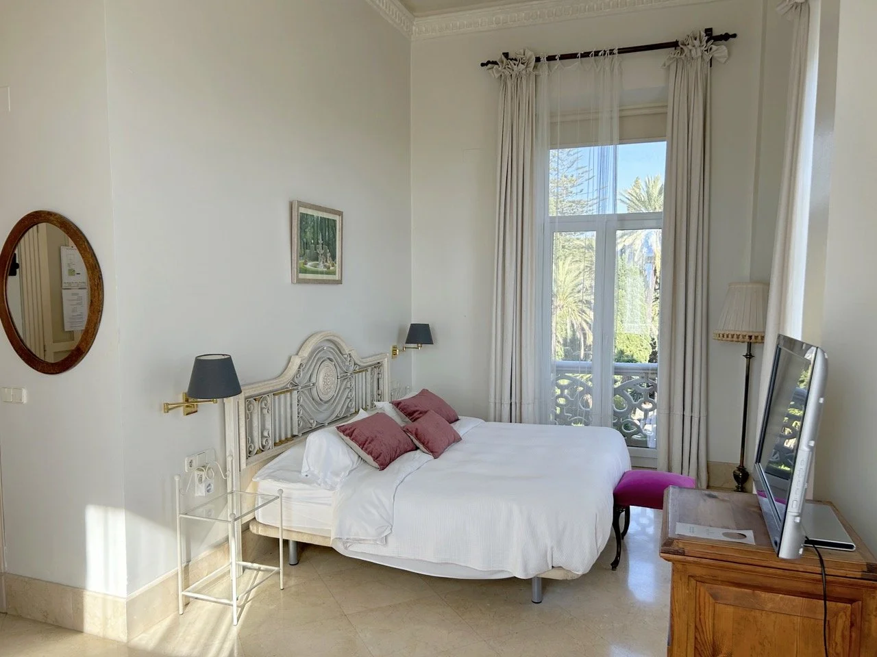A bedroom with white walls and curtains, a bed with white bedding and purple pillows, a wooden dresser with a television, a pink cushioned bench, and a large window showing trees and a blue sky outside.