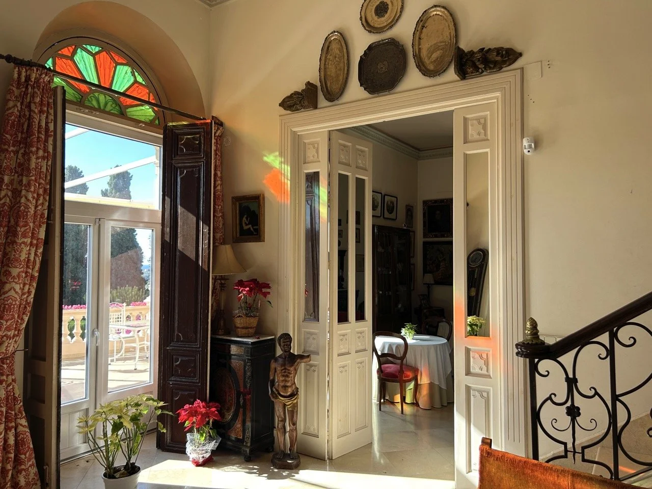 Interior of a vintage-style house with stained glass on the window above a sliding door, patterned curtains, indoor plants, antique furniture, and decorative plates and artifacts on the wall. Sunlight creates colorful reflections inside.