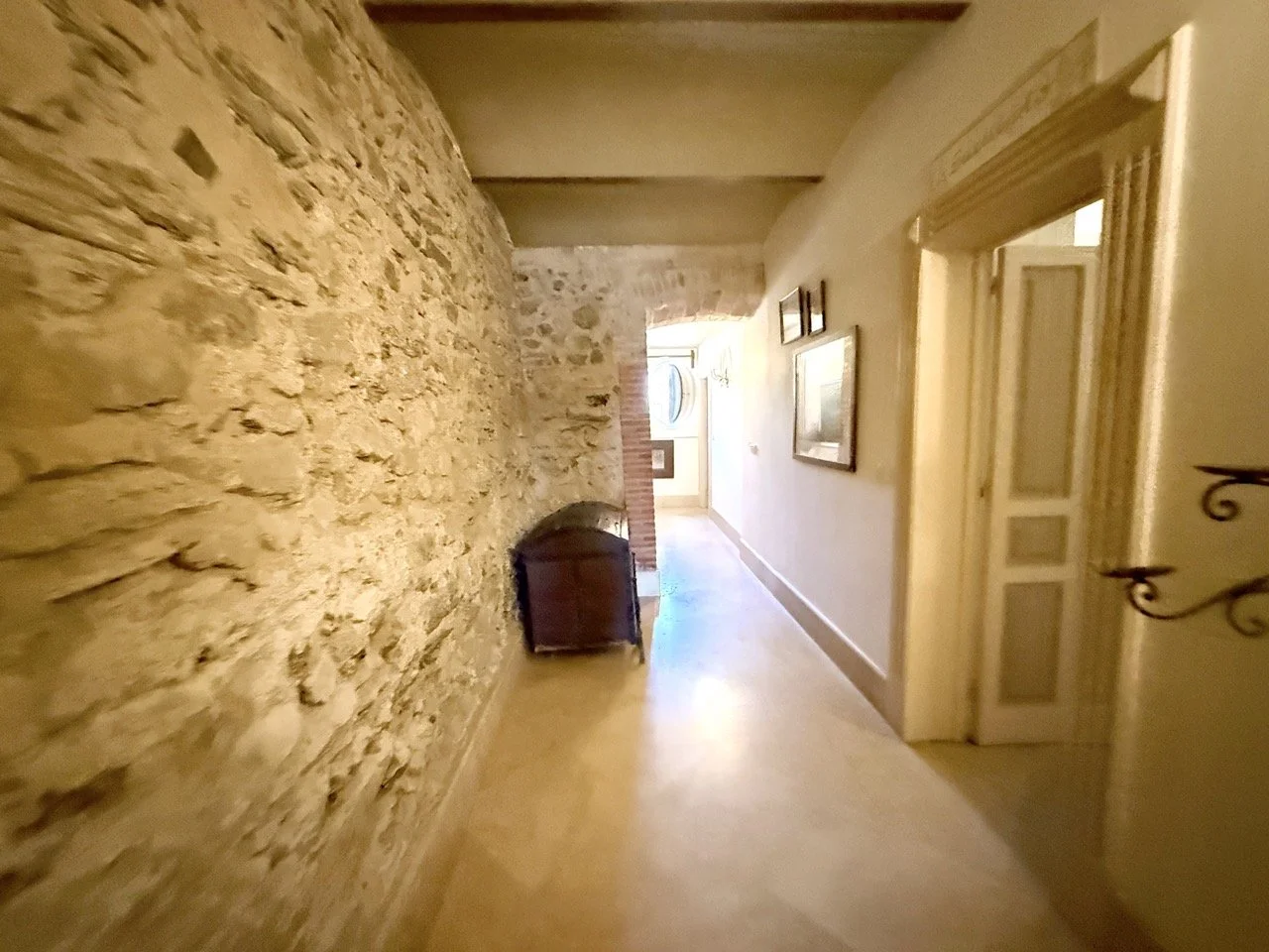 A hallway with an exposed stone wall on the left and a white wall with framed pictures on the right. There is a wooden chest against the stone wall. The hallway leads to a door with sunlight shining through. The floor is light-colored wood or laminat