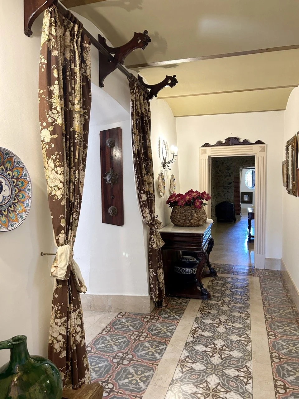 Elegant hallway with patterned tile flooring, floral curtains hung on decorative wooden rods, and vintage wall decor. A wooden table with a basket of pink flowers is against the wall, leading to a room with a brick wall and a mirror visible in the ba