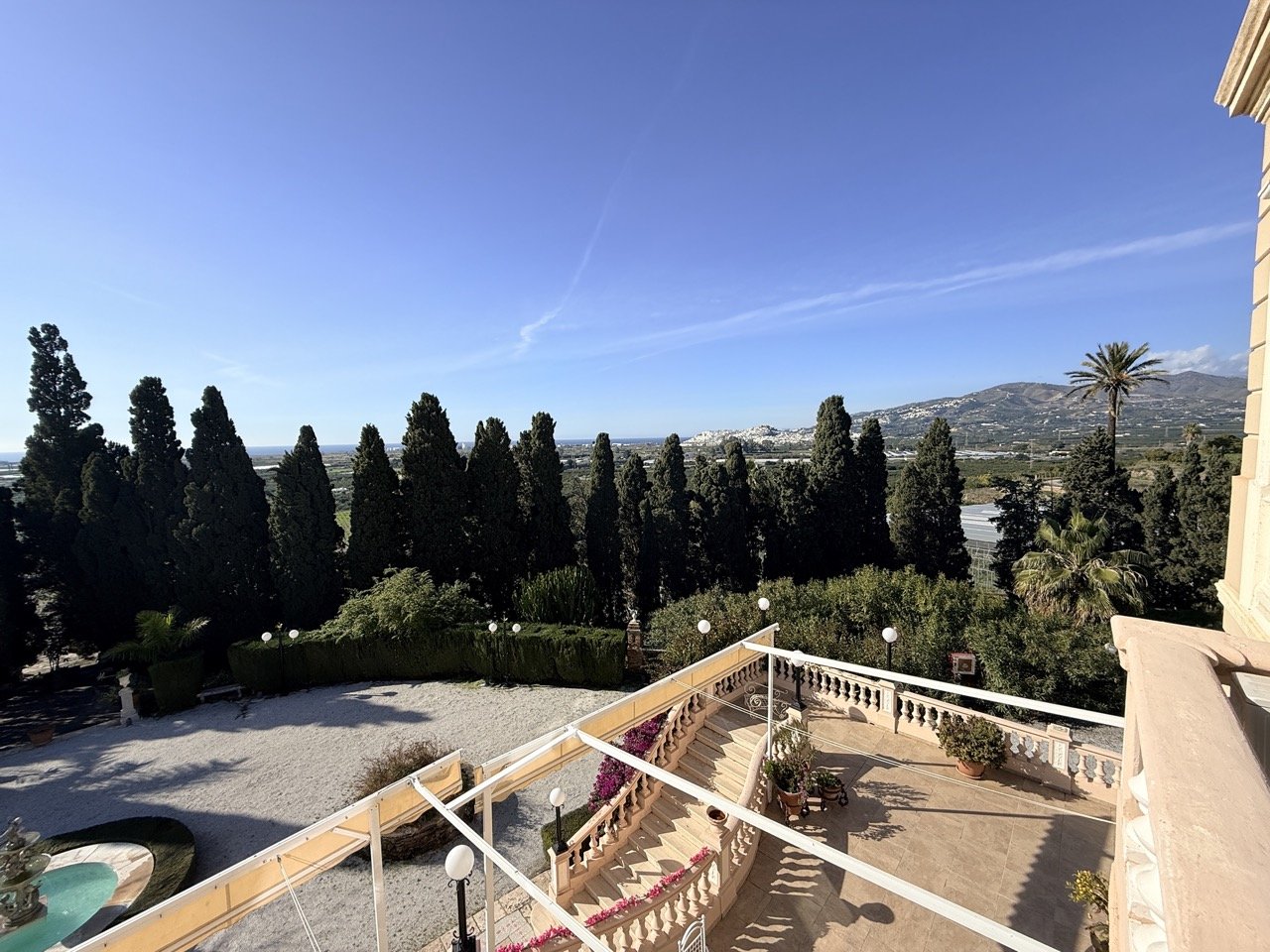 View from a balcony overlooking a landscaped garden with trees, some palm trees, and distant mountains, under a clear blue sky.