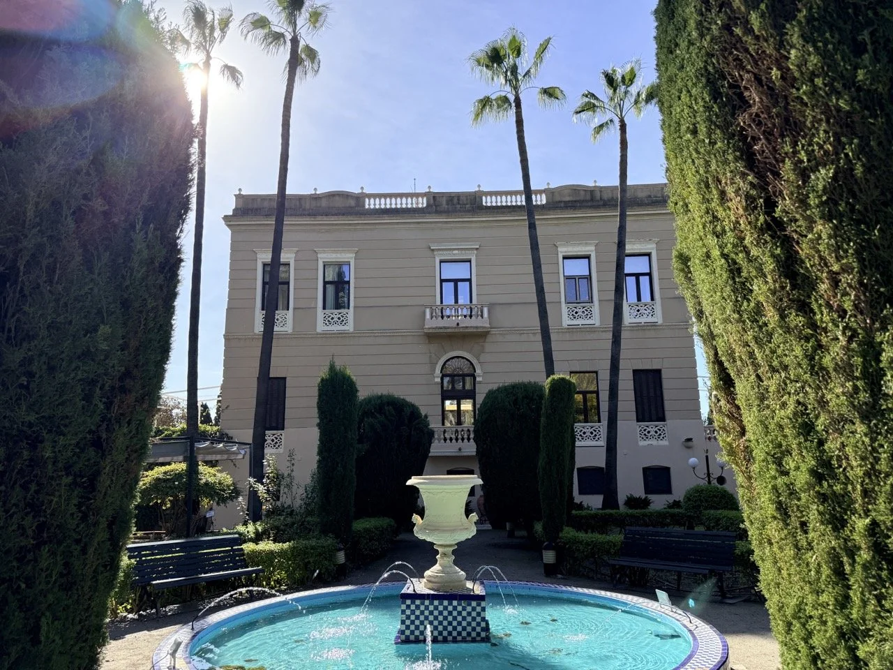 A luxurious residential building with four stories, featuring a central balcony, large windows, and decorative railings, surrounded by tall palm trees, trimmed bushes, and a fountain in the foreground, with sunlight filtering through the trees.