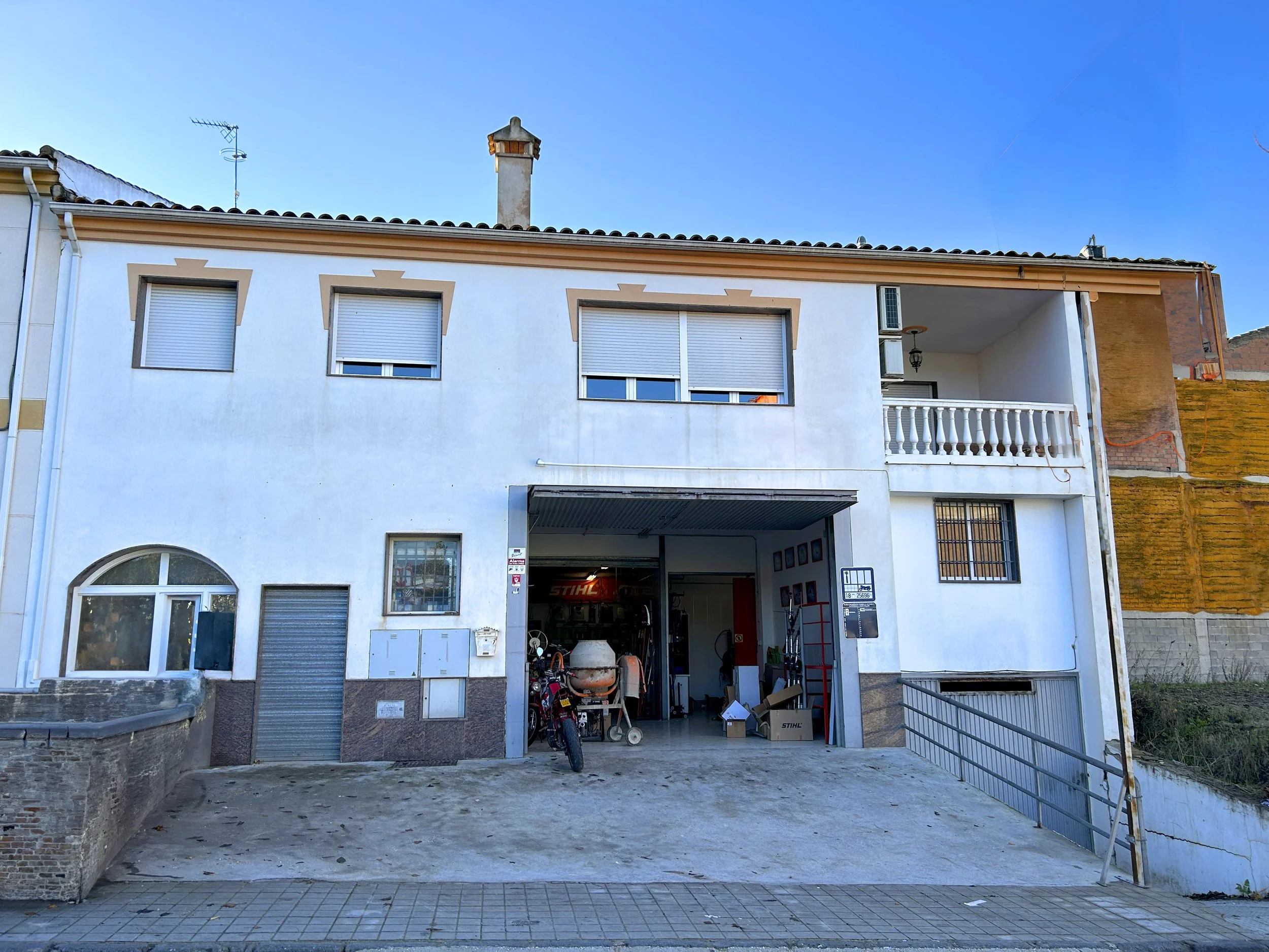 White multi-story building with a garage on the ground floor, parked motorcycle, and storage boxes; three upper windows with closed shutters, small balcony with balustrade on the right side, and a chimney on the roof.