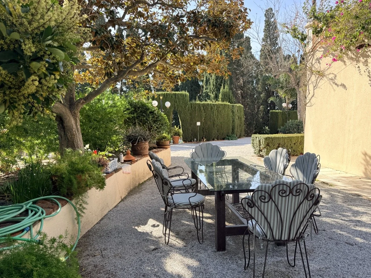 Outdoor patio with a black glass table surrounded by vintage-style metal chairs with striped cushions, lush green trees, trim hedges, potted plants, and gravel ground under a sunny sky.