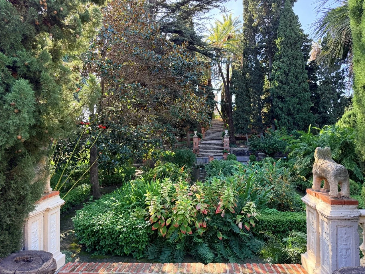 A lush garden with a variety of trees, bushes, and plants, featuring a stone statue of an animal on a pedestal on the right, a decorative staircase in the background, and potted trees on either side.
