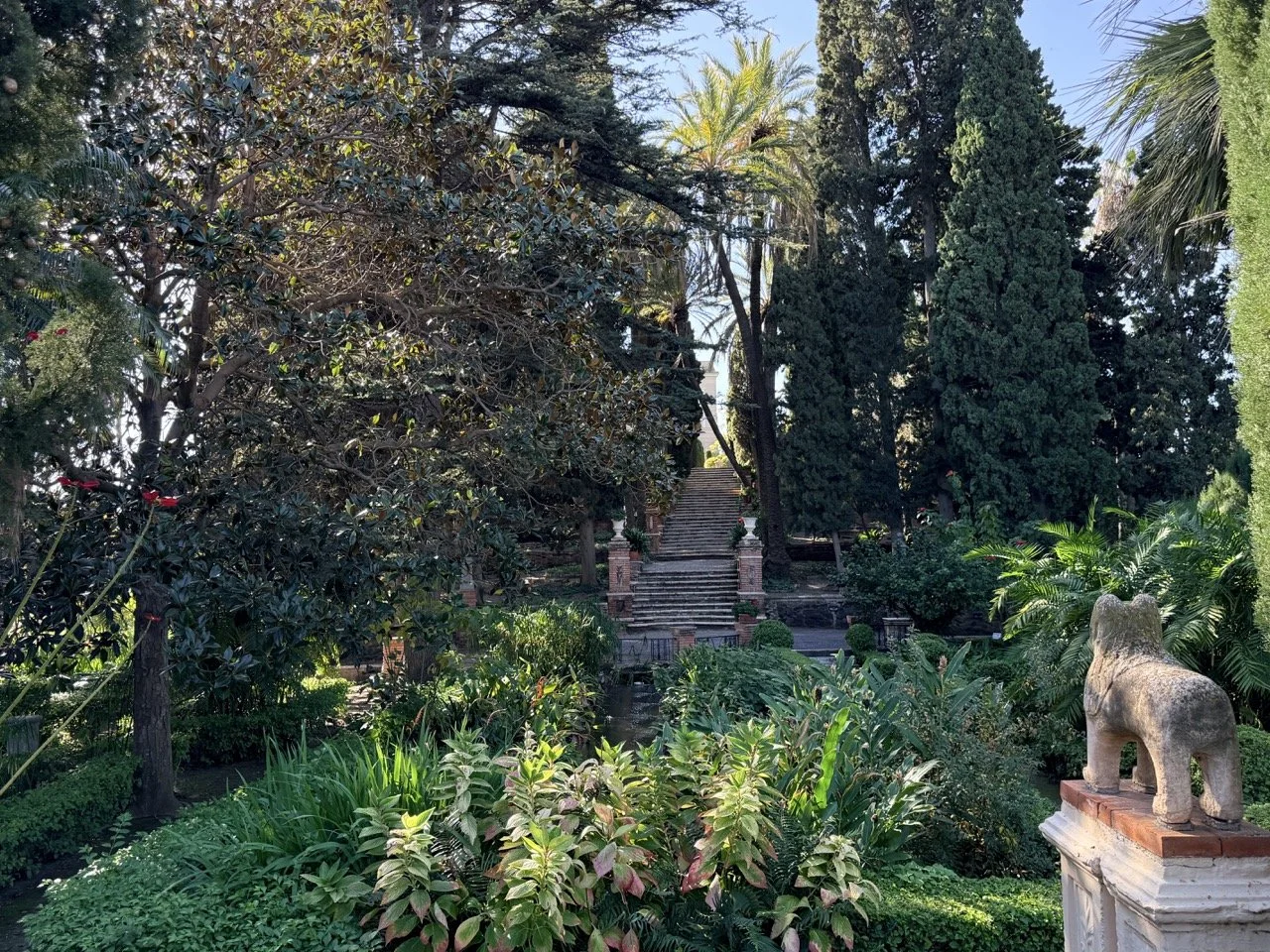 A lush garden with stairs leading up to a building, surrounded by tall trees, including palms and cypress. A stone lion statue is positioned on a pedestal in the foreground.