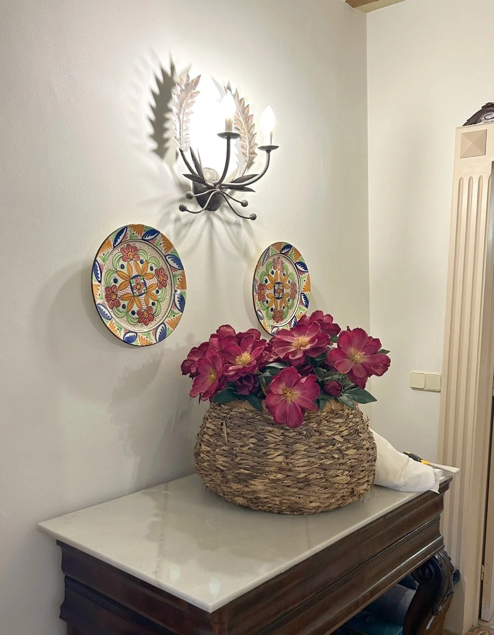 A decorated indoor wall with a vintage-style wall sconce light fixture casting shadows. Below are two colorful, patterned ceramic plates hung on the wall. On a wooden table with a white marble top, there is a large woven basket filled with pink flowe