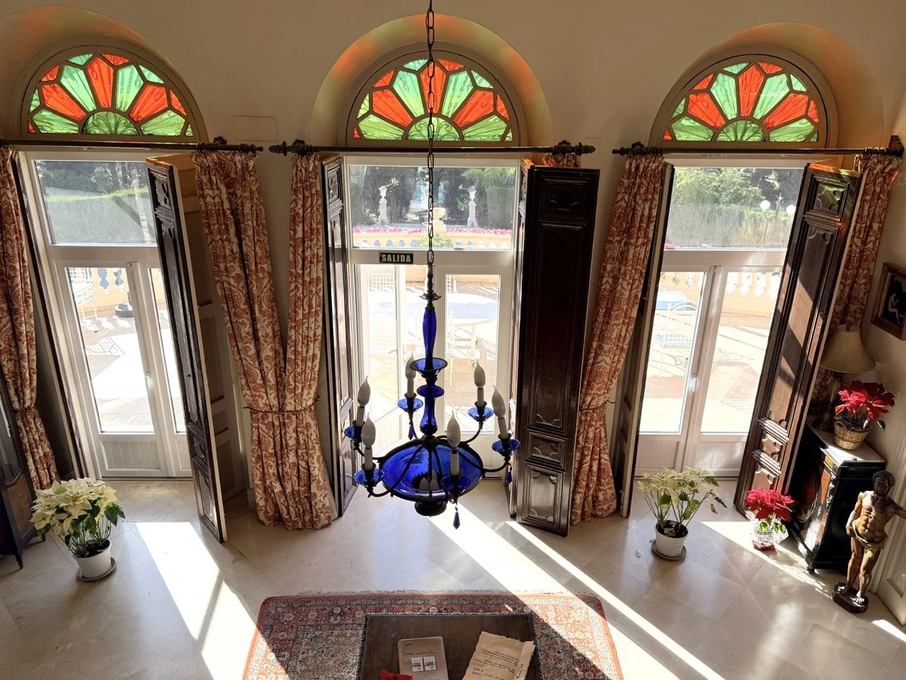 An interior view of a room with large windows and stained glass arches at the top, curtains on each side, a blue chandelier hanging from the ceiling, potted plants near the doors, and a rug on the floor with a table on it.