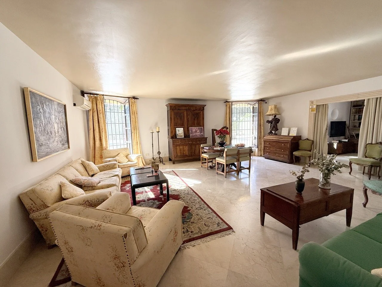 Spacious living room with cream-colored sofas, a green armchair, wooden coffee tables, and vintage decor, illuminated by natural light from large windows with gold curtains.