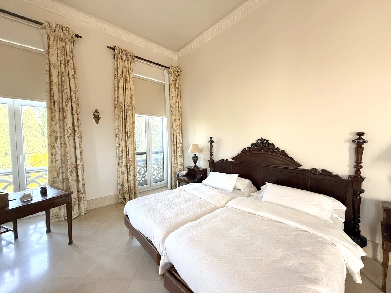 A hotel room with two twin beds with dark wooden headboards, flanked by a nightstand with a lamp, and light-colored walls. There are large windows with floral curtains, a small wooden table near the window, and a balcony visible through the window.