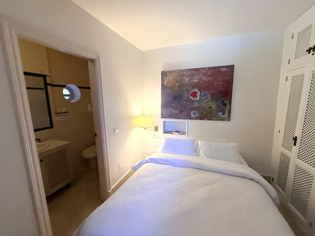 A cozy hotel bedroom with a white bed, a small bedside lamp, colorful abstract painting on the wall, and an adjoining bathroom visible through an open door.