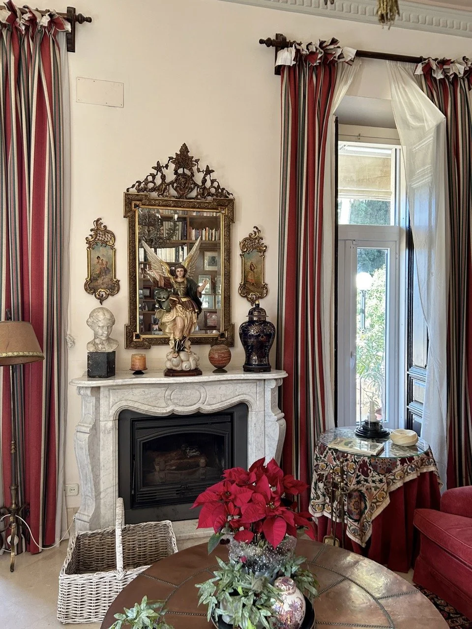 Living room with a white marble fireplace, reflection of a bookshelf in the mirror, a religious angel statue on the mantel, framed artwork, and decorative objects. Red and beige striped curtains around a window, poinsettia plant on a wooden table, an