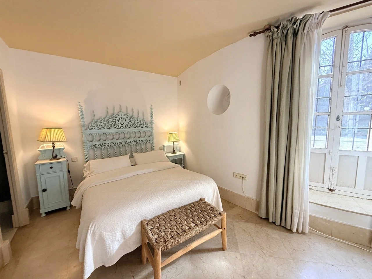 Cozy bedroom with a carved light blue headboard, white bedding, two bedside lamps, beige curtains, a window showing trees outside, and a small woven bench at the foot of the bed.