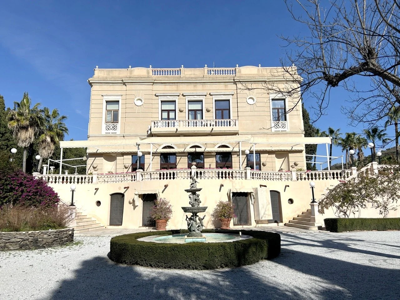 A large, elegant beige mansion with multiple balconies and decorative architectural details, surrounded by a garden with trees, bushes, a fountain, and gravel pathways under a clear blue sky.