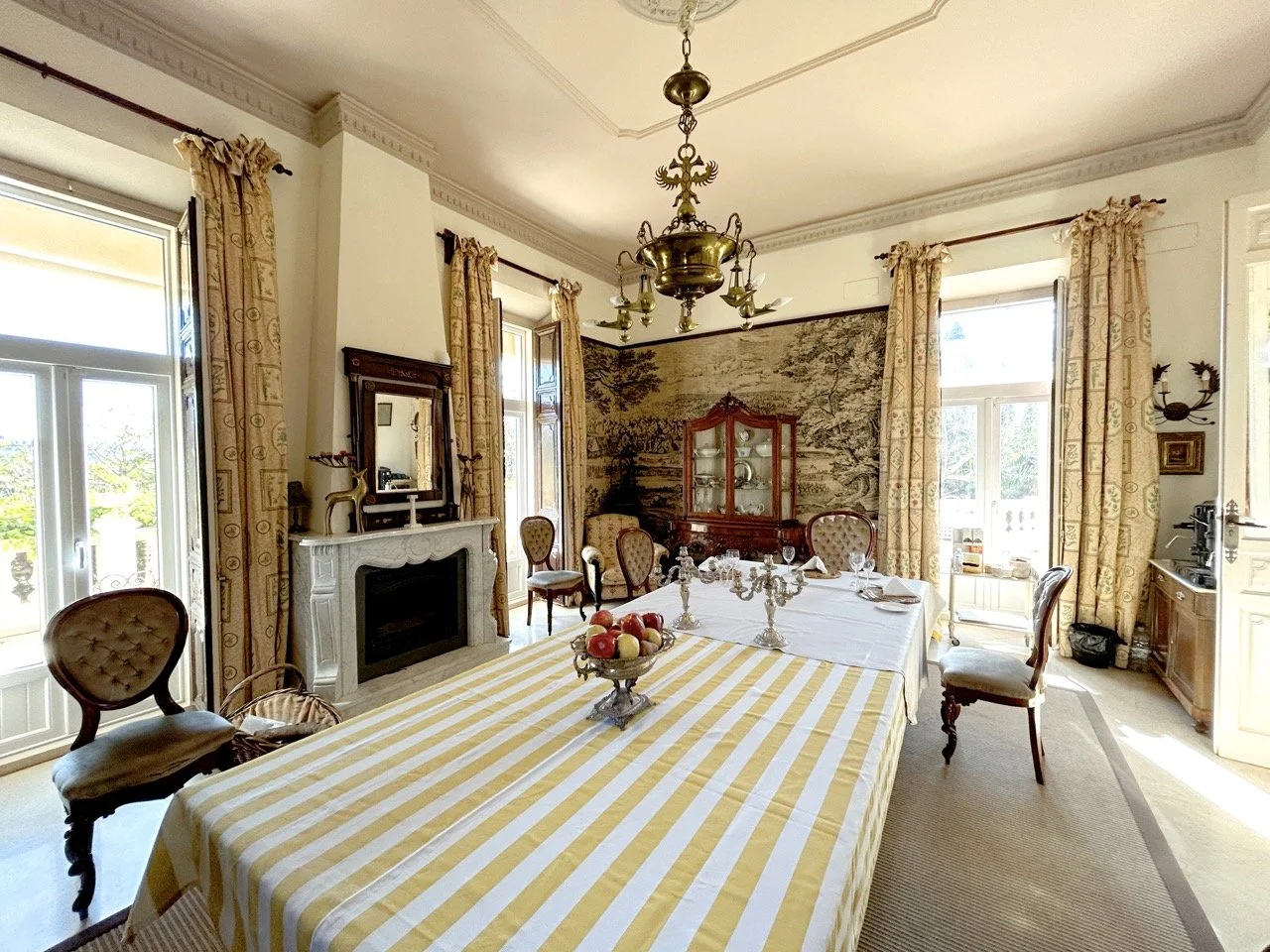 Elegant dining room with a long table covered in a yellow and white striped tablecloth, set with silver candlesticks, surrounded by wooden chairs with upholstered seats. There are large windows with patterned curtains, a vintage chandelier hanging fr