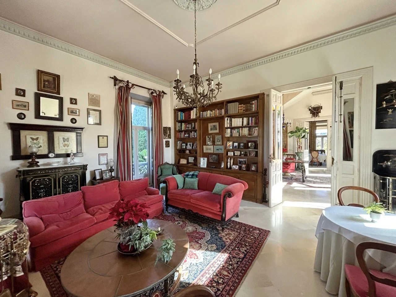 A living room with red sofas, a wooden bookshelf filled with books and photos, a chandelier, and a window with curtains. There is also a dining table with a white tablecloth and a small round table with a poinsettia plant.