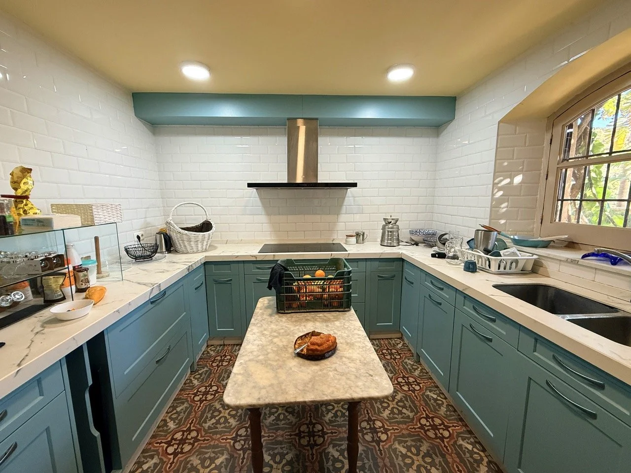Cozy kitchen with teal cabinets, white brick walls, and a central marble table. Includes a window with bars, a range hood, and various dishes and kitchen items on the countertops.