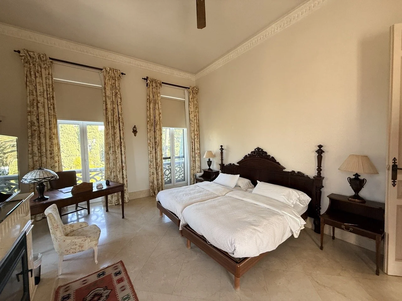 A bedroom with two beds, carved dark wood headboards, bedside tables with lamps, and floral curtains, brightened by natural light from windows and balcony doors.
