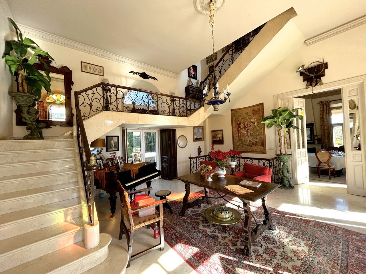 Spacious living room with staircase, ornate furniture, potted plants, decorative paintings, and sunlight streaming in through open doors.