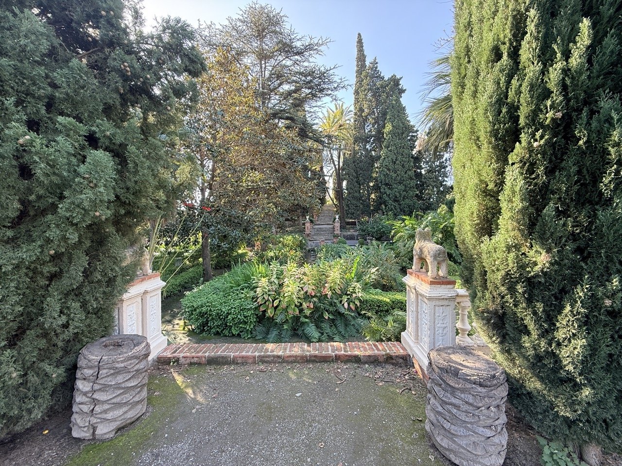 A garden entrance framed by tall, lush, green trees and bushes, with decorative stone and brick elements, including a statue of a lion on a pedestal.
