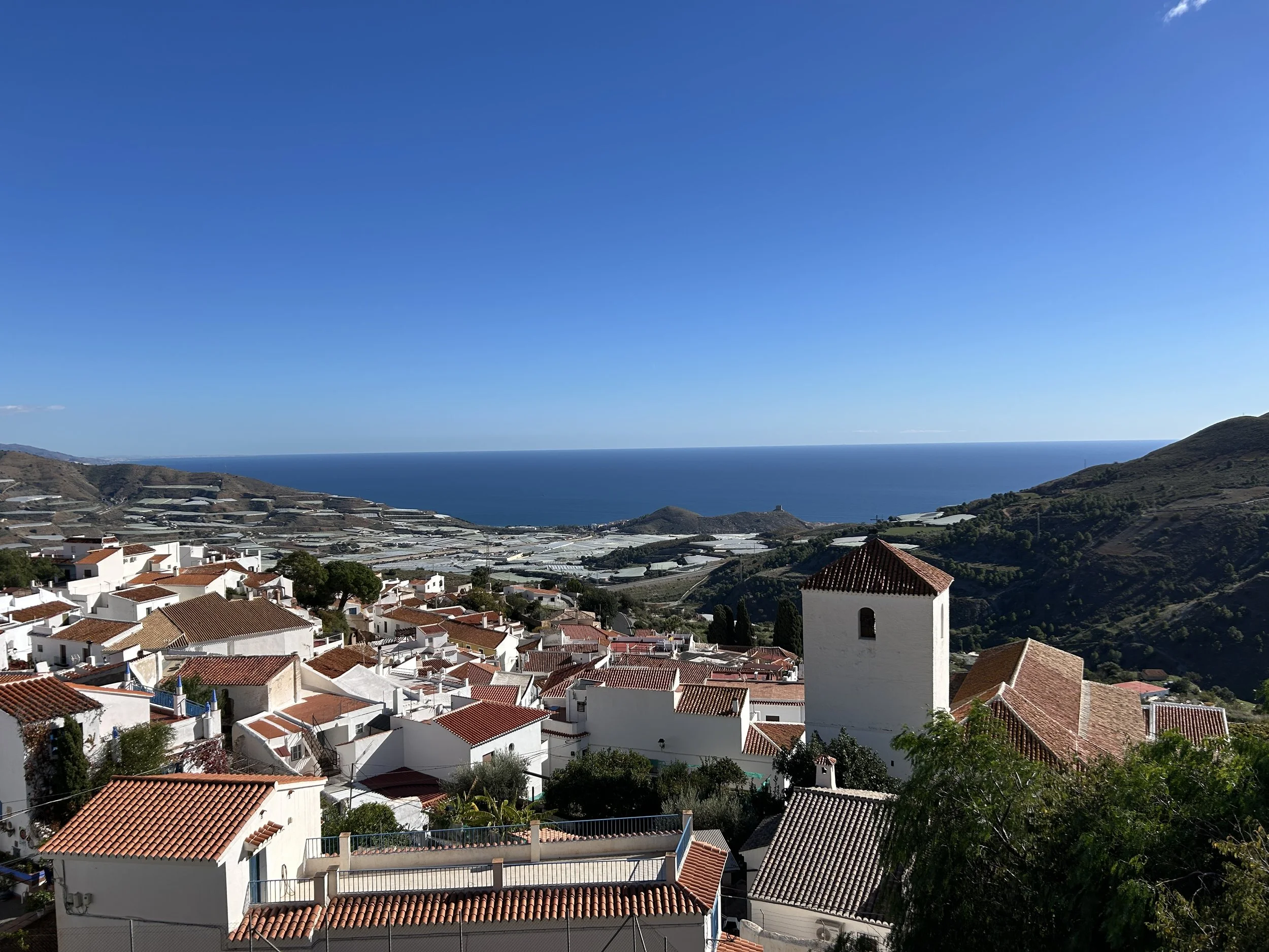 A coastal town with white buildings and red-tiled roofs, overlooking the ocean and surrounding hills, under a bright blue sky.