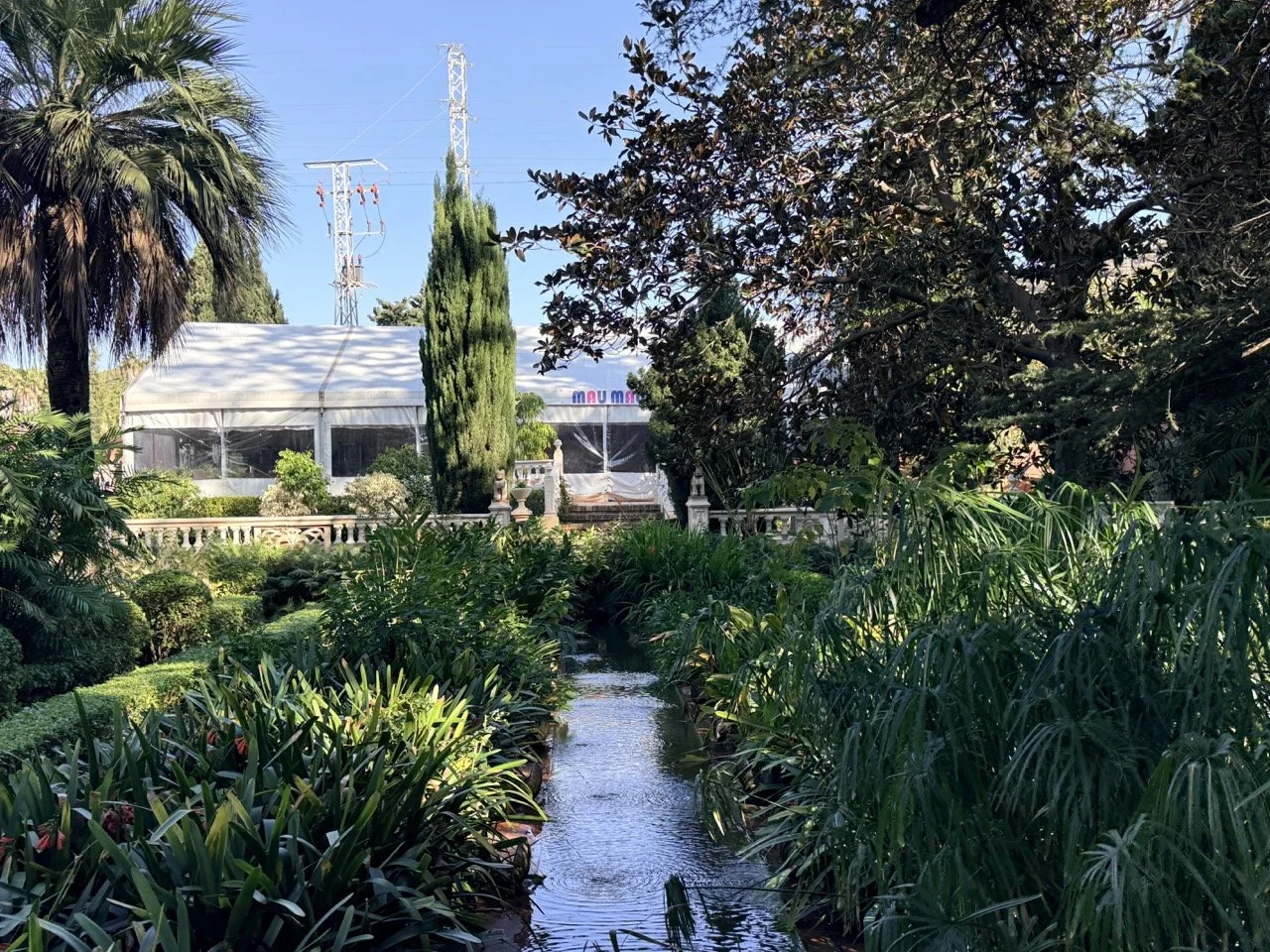 A lush garden with a small stream running through it, surrounded by green plants and trees. In the background, there is a white tent and a sign that reads 'MUM' or 'NAUM'.