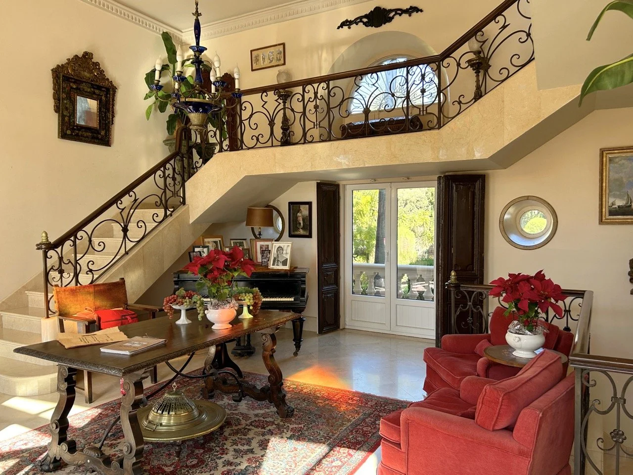 Living room with a staircase, a chandelier, a grand piano, a wooden table with poinsettia plant, red armchair, and potted plants, decorated with paintings and framed photos.